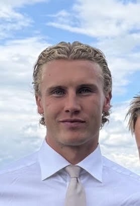Professional headshot of a man with blonde hair wearing a light blue shirt and tie against a cloudy sky background